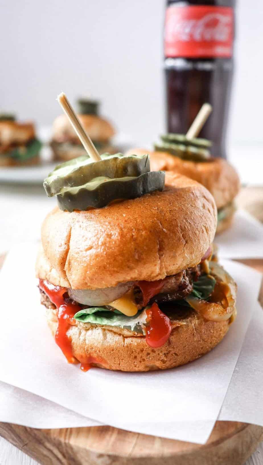 two burgers on a wooden board with a coke in the background. 