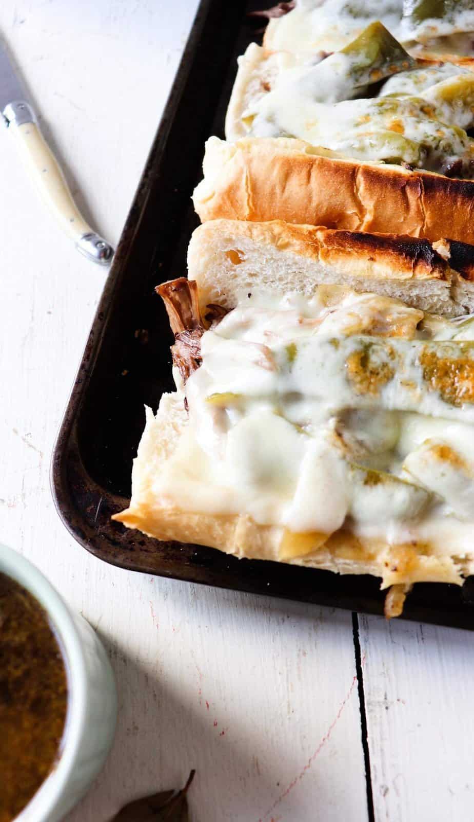 two completed beef sandwiches on a dark baking sheet with a knife and cup of au jus to the side