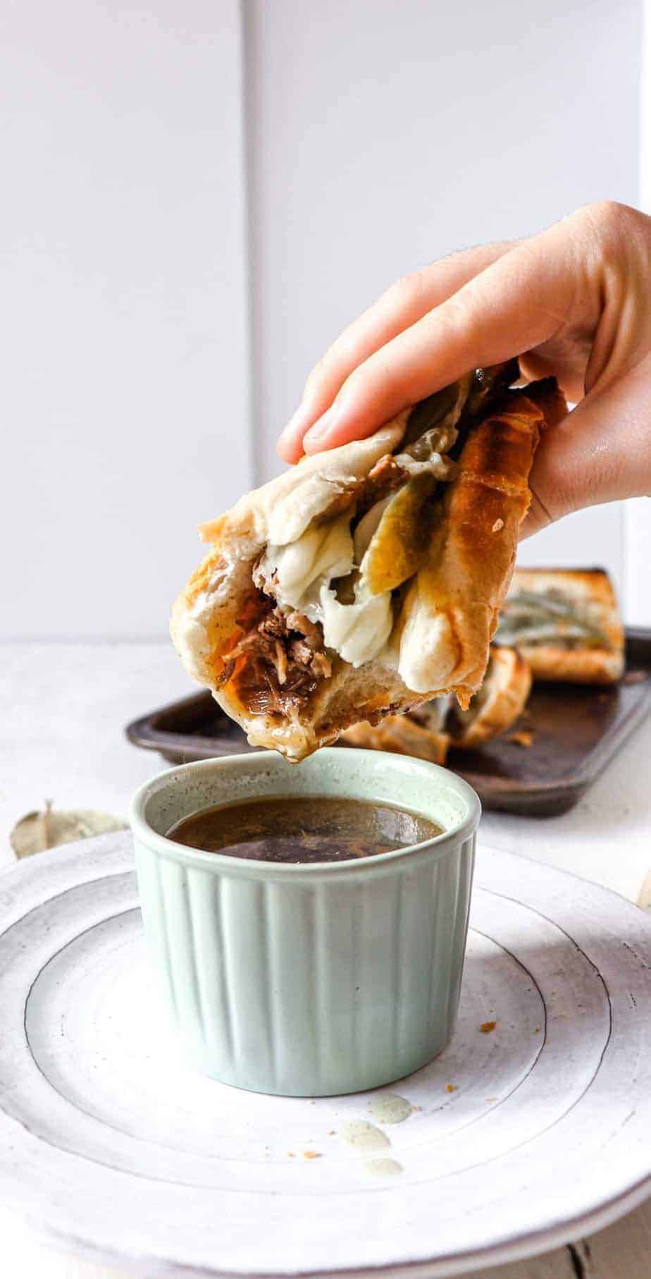 hand holding a beef sandwich that has a bite taken out of it and is being dipped into a ramekin of au jus.