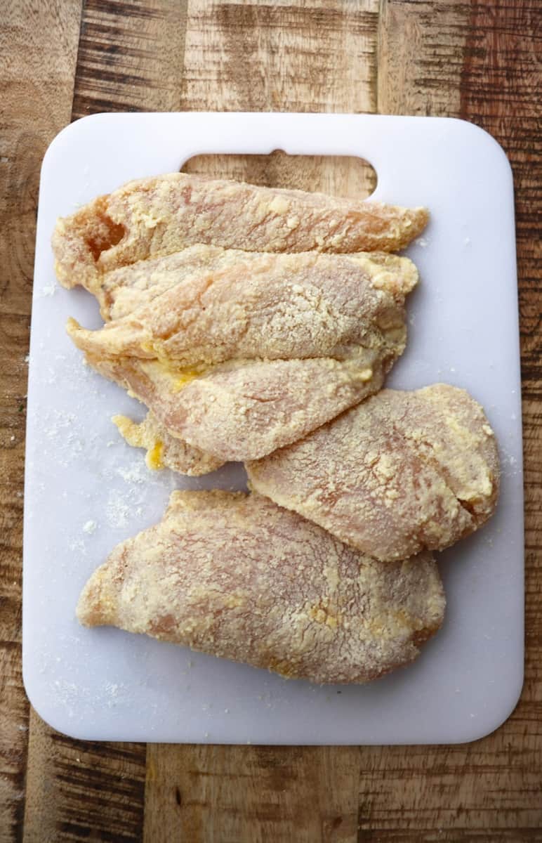 chicken breasts coated in parmescan on a white cutting board.