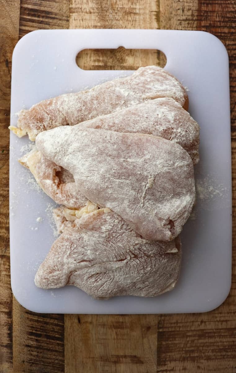 chicken breasts coated in flour on a white cutting board.
