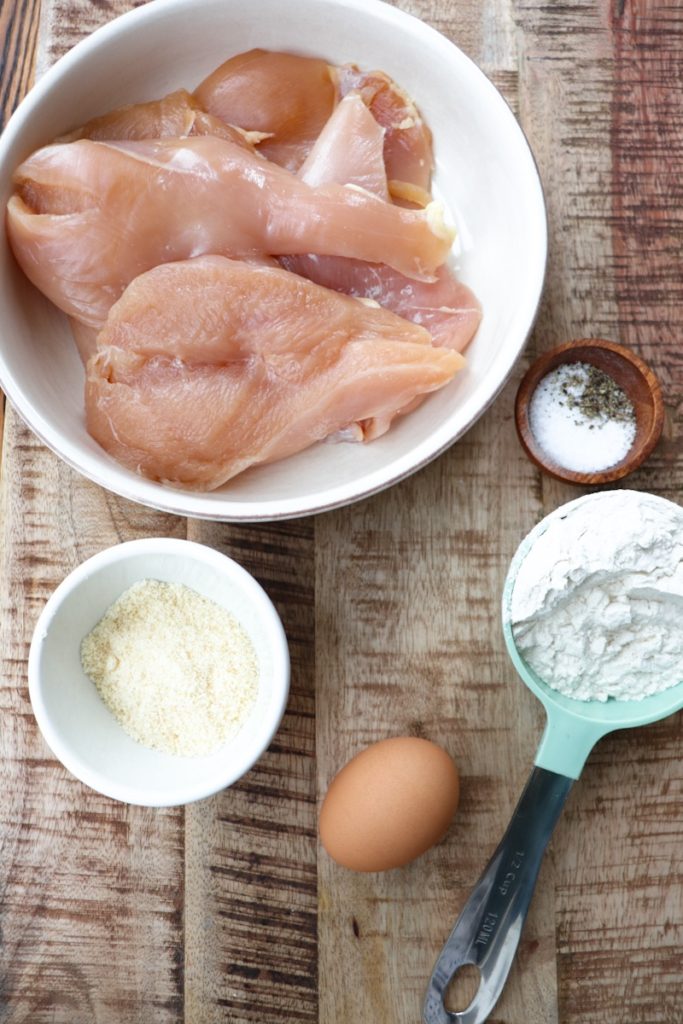 raw chicken breasts, salt, pepper, flour, parmesan cheese, and an egg measured out on a wooden cutting board