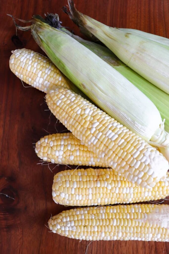 white and yellow corn on the cob both in and out of husk on a dark wooden background