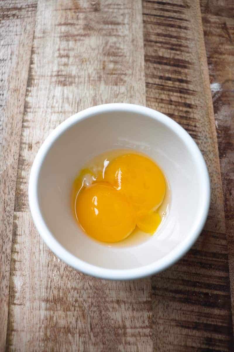 two egg yolks in a white bowl on a light wood background
