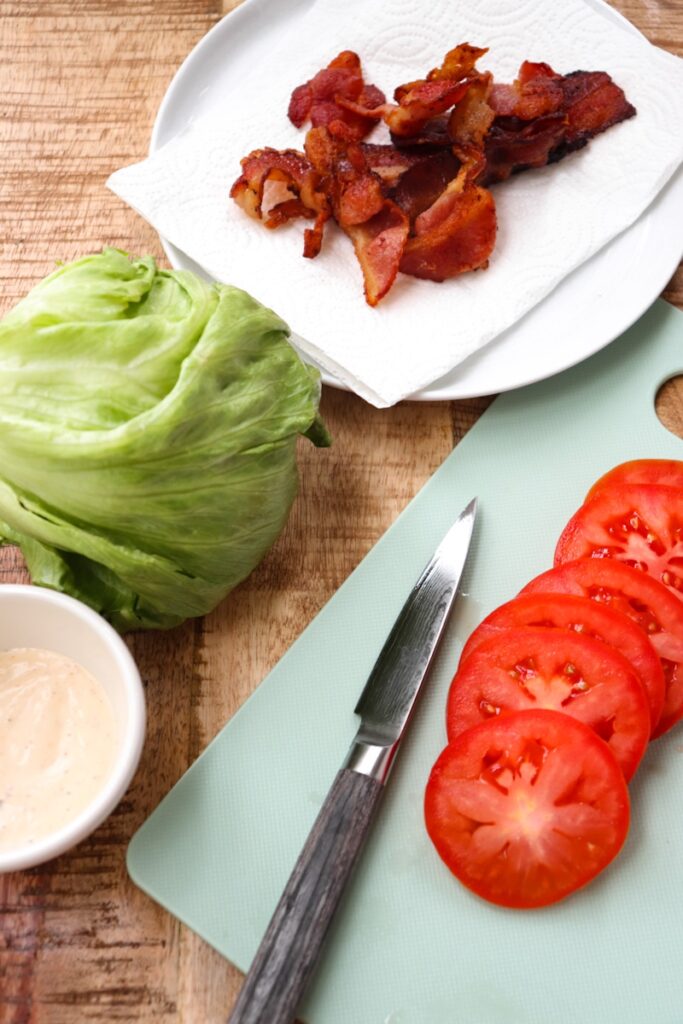 tomatoes sliced on a mint green cutting board with a knife next to it, ranch in a small white bowl, iceberg lettuce, and bacon on a paper towel on a white plate