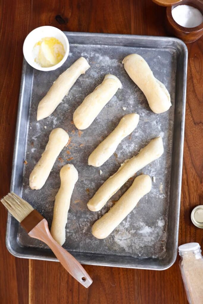 eight cooked sourdough breadsticks on a silver baking sheet with melted butter, a pastry brush, garlic powder, and salt around it