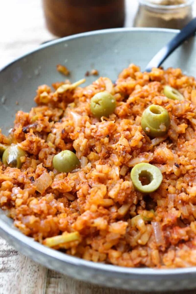 angled shot of red spanish rice with sliced green olives on top in a shallow grey bowl