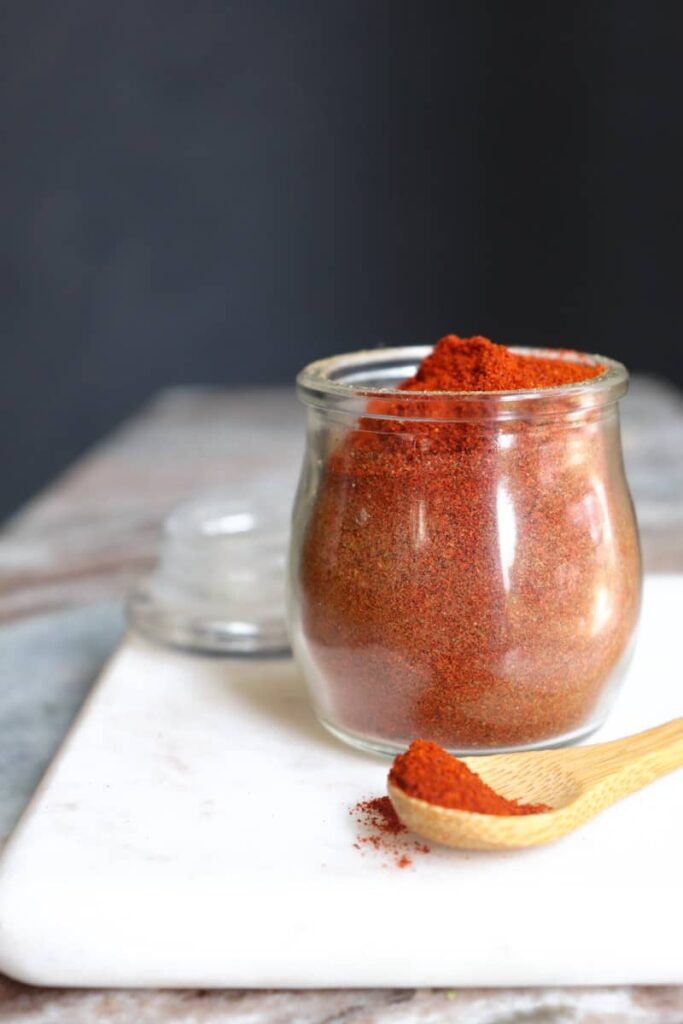 chili powder in a glass jar with a wooden spoon placed at an angle in front on a white marble cutting board