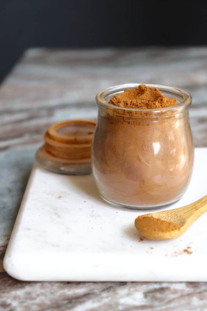 ground cinnamon in a glass jar with a wooden spoon placed at an angle in front on a white marble cutting board