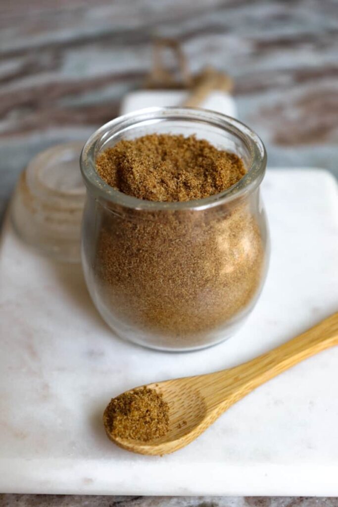 ground cumin in a glass jar with a wooden spoon placed at an angle in front on a white marble cutting board