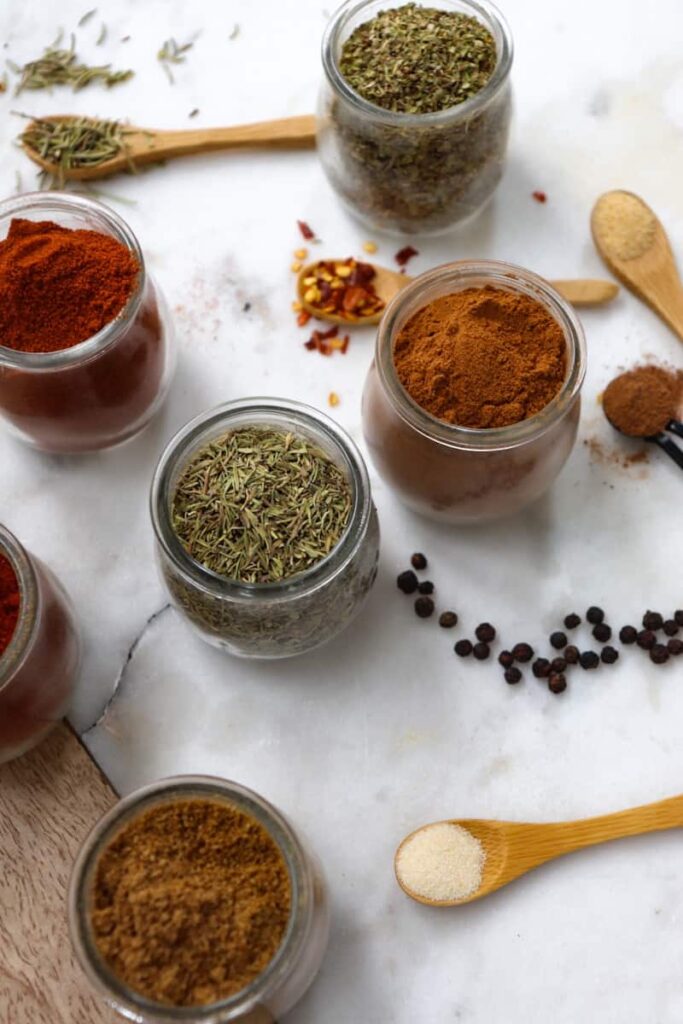 kitchen spices and herbs in glass jars and on wooden spoons placed decoratively on a whtie marble slab