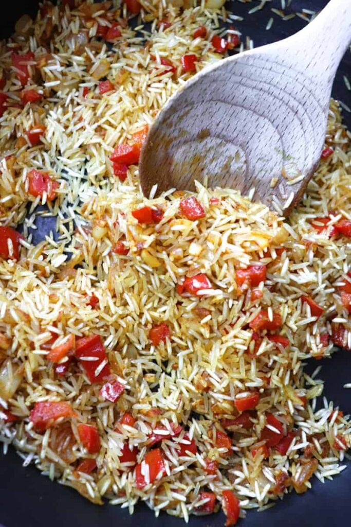 dry rice, diced peppers and onions, and spices in a dark grey pan with a wooden spoon coming out
