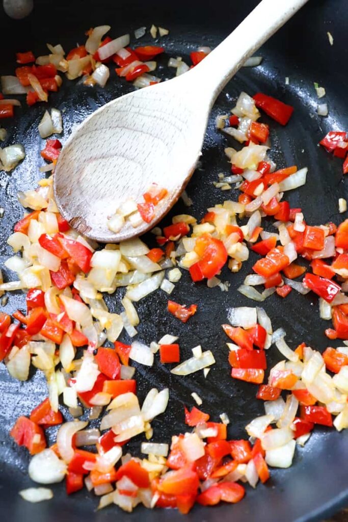 sauteing onions, garlic, and red bell pepper in a dark gray pan with a wooden spoon  