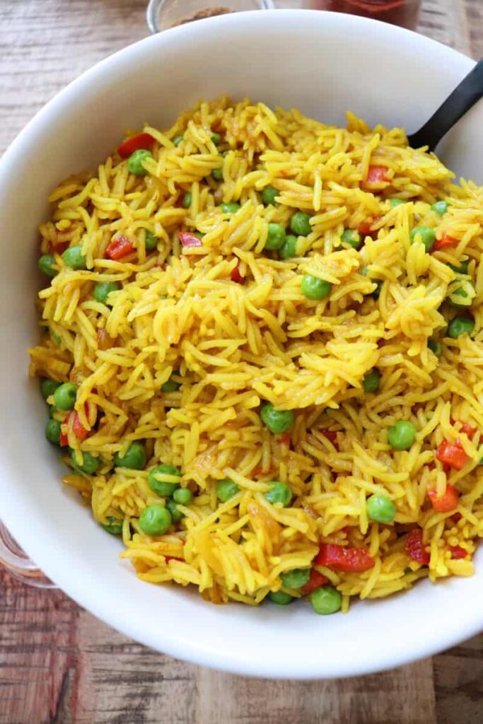 yellow rice in a white bowl with peas and diced red pepper. two spice jars open on the wooden background