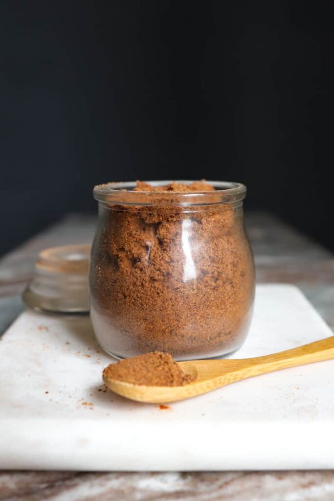 ground nutmeg in a glass jar with a wooden spoon placed at an angle in front on a white marble cutting board