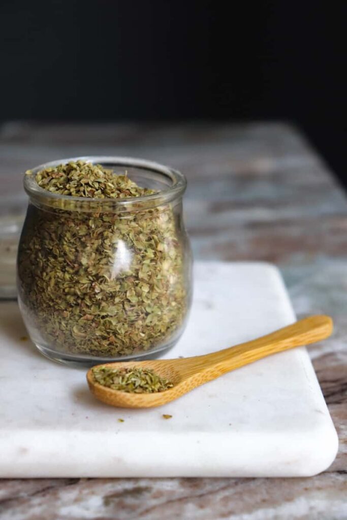 dried oregano in a glass jar with a wooden spoon placed at an angle in front on a white marble cutting board