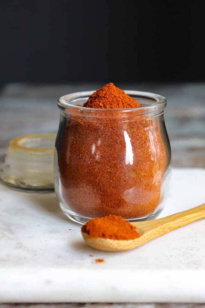 smoked paprika in a glass jar with a wooden spoon placed at an angle in front on a white marble cutting board