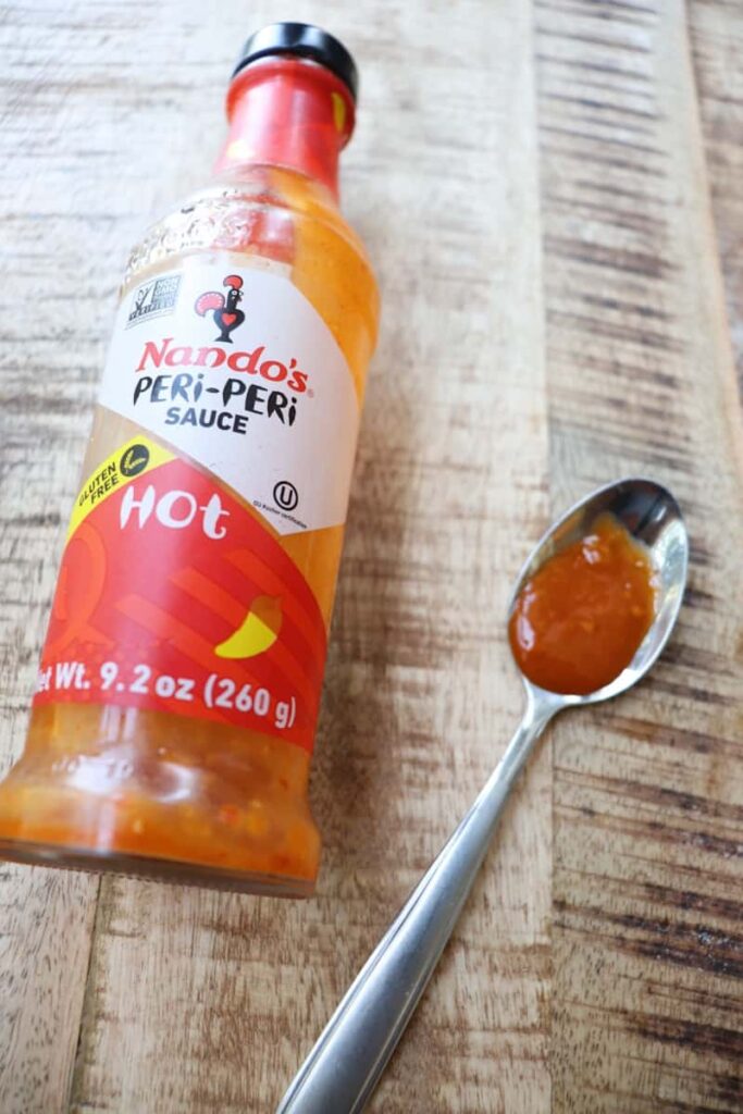 glass jar of nandos medium hot sauce laying down next to a silver spoon with hot sauce on it.