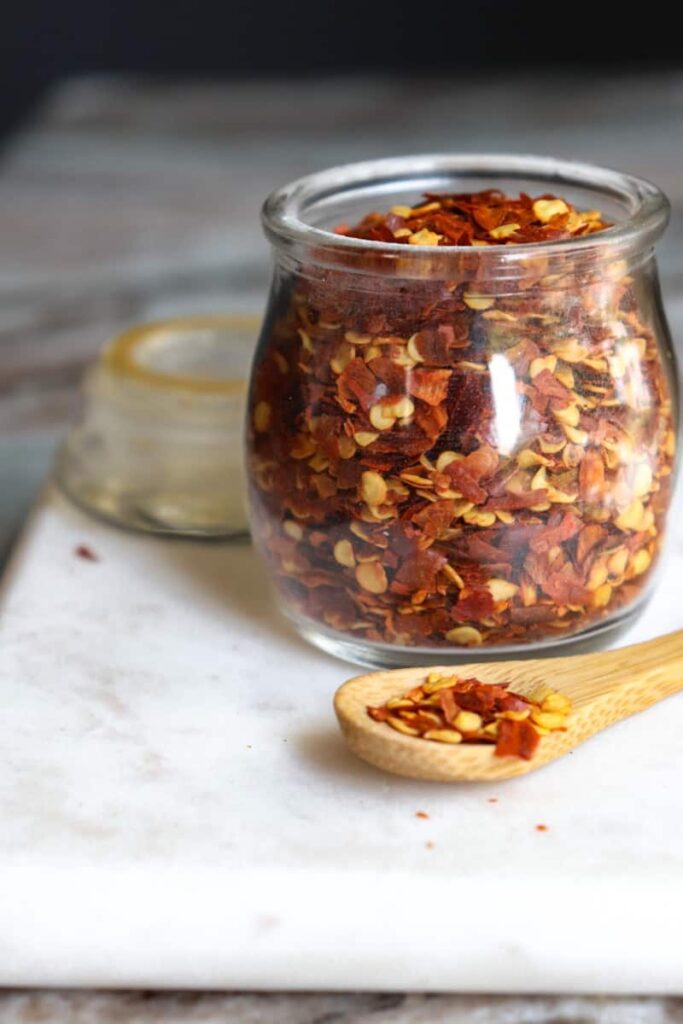 red pepper flakes in a glass jar with a spoon in front of them.