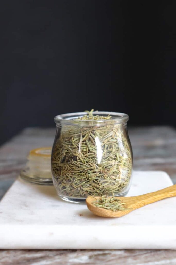 dried rosemary in a glass jar with a wooden spoon placed at an angle in front on a white marble cutting board