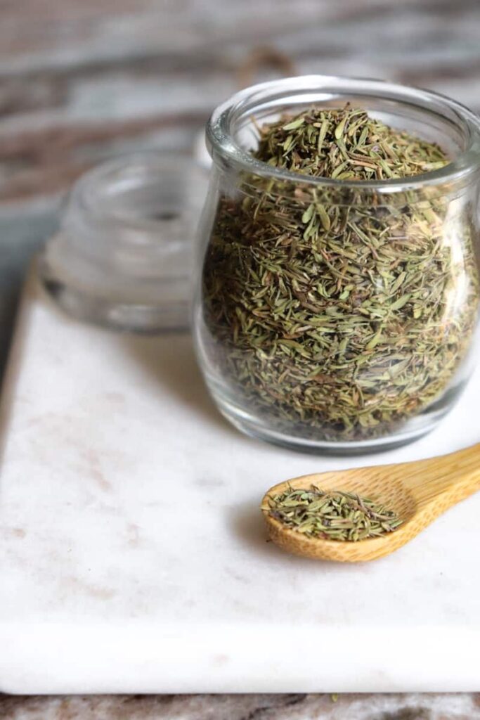 dried thyme in a glass jar with a wooden spoon placed at an angle in front on a white marble cutting board