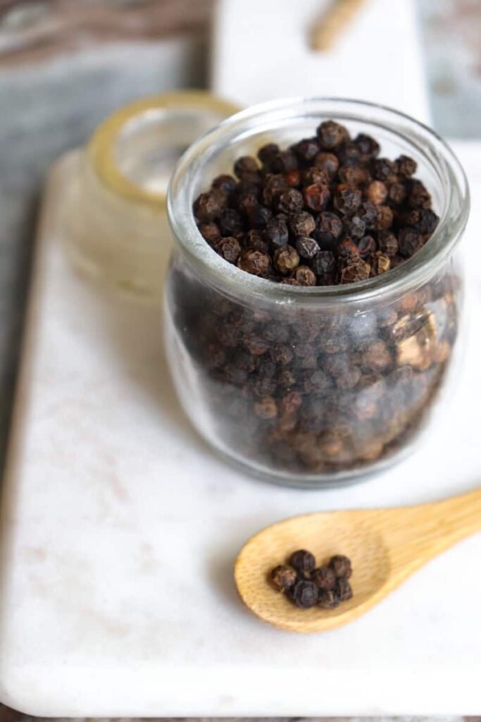 whole peppercorns in a glass jar with a wooden spoon placed at an angle in front on a white marble cutting board