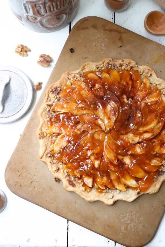 apple tart on a stone baking dish with walnuts and jars scattered on the side