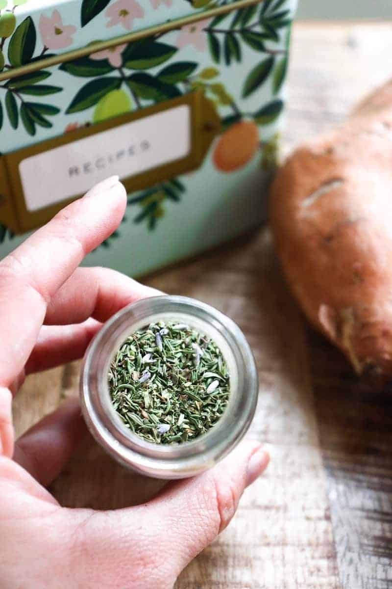 hand holding glass jar of herbs de provence in front of a recipe tin.