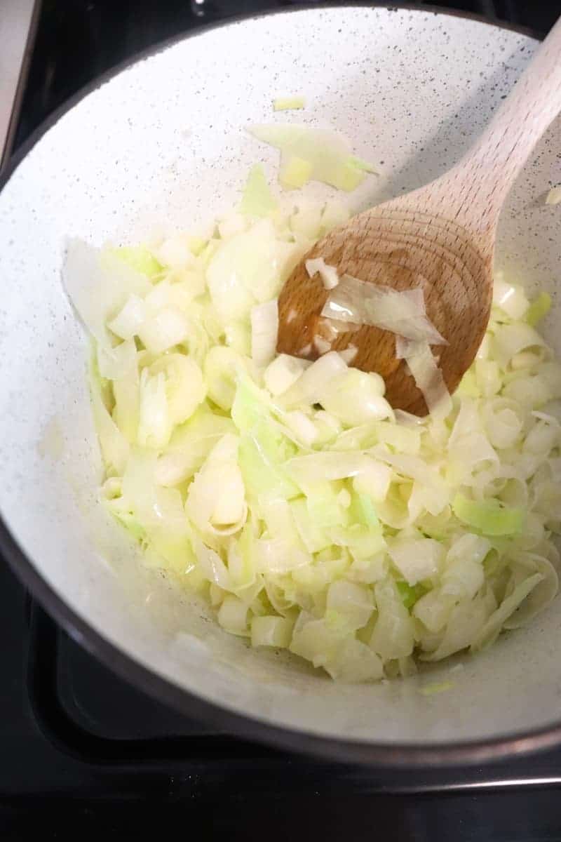 leeks and garlic sauteing in a white speckled pot with a wooden spoon to stir