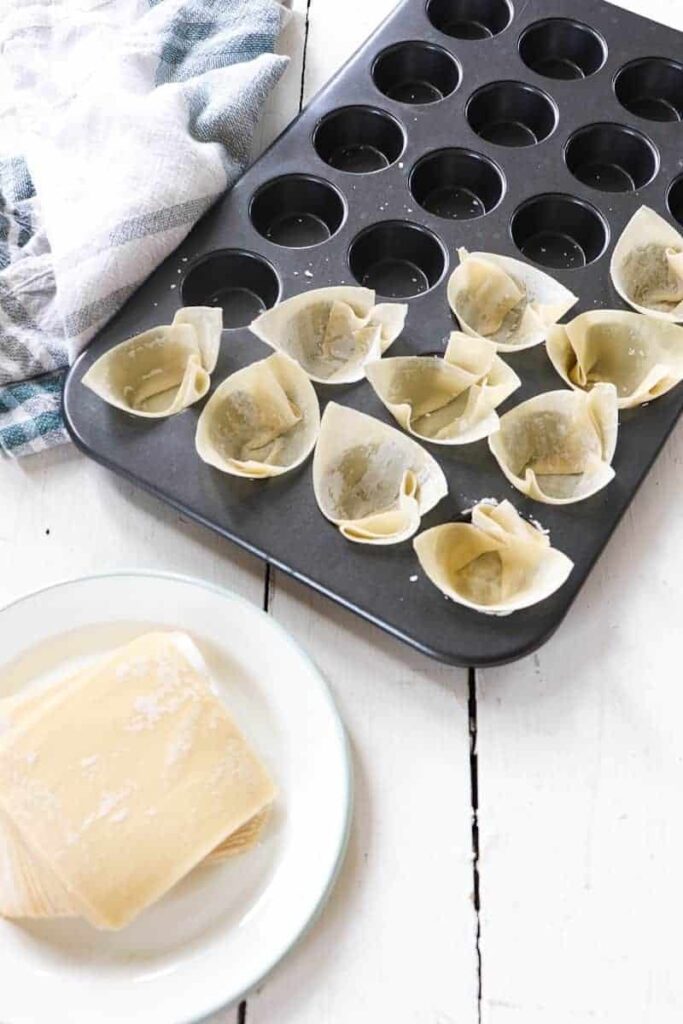 wonton wrappers being placed in a mini muffin tin on a white table with a striped towel to the side