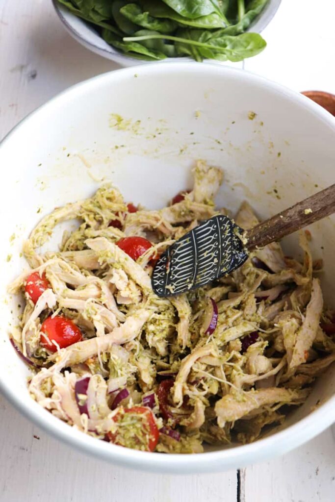 pesto chicken salad ingredients mixed in a white bowl with a spatula