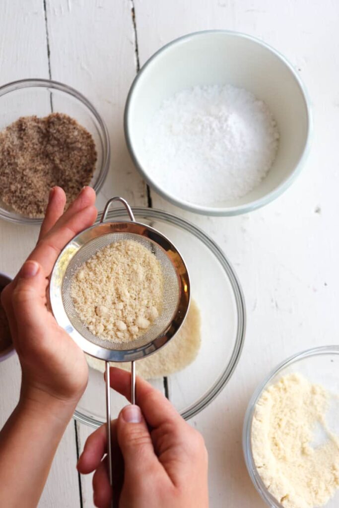 hand sifting almond flour into a glass bowl with bowls of other ingredients scattered around