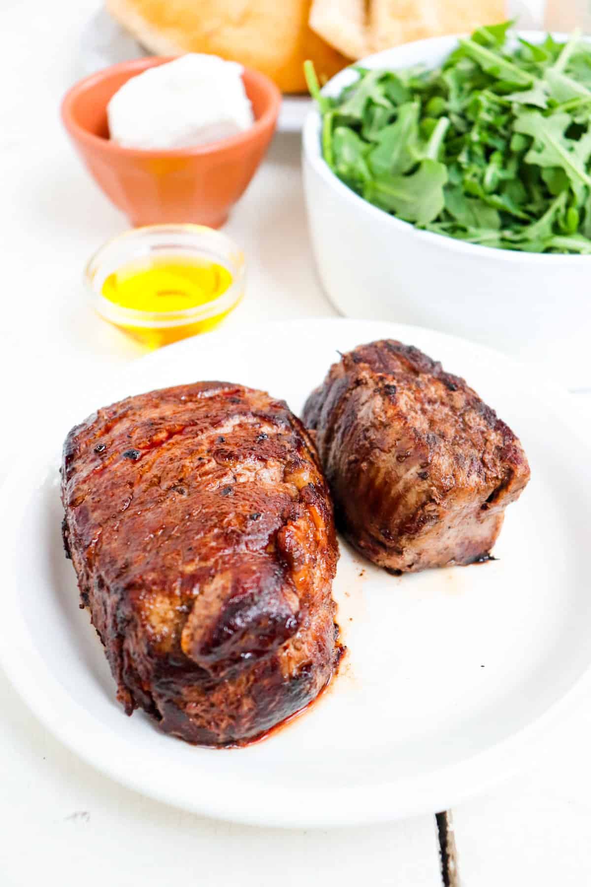 two cooked sirloins on a plate with additional ingredients in the background.