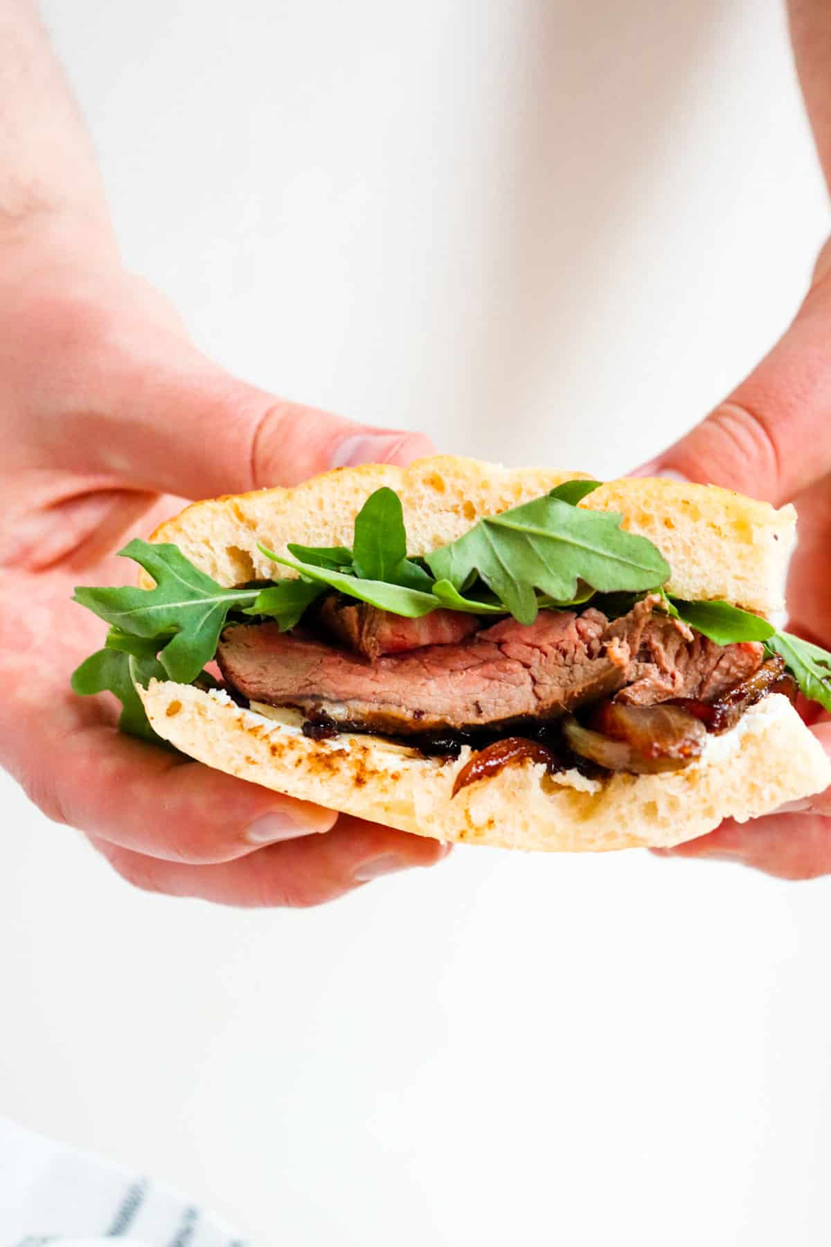 hands holding a steak sandwich with arugula leaves poking out.