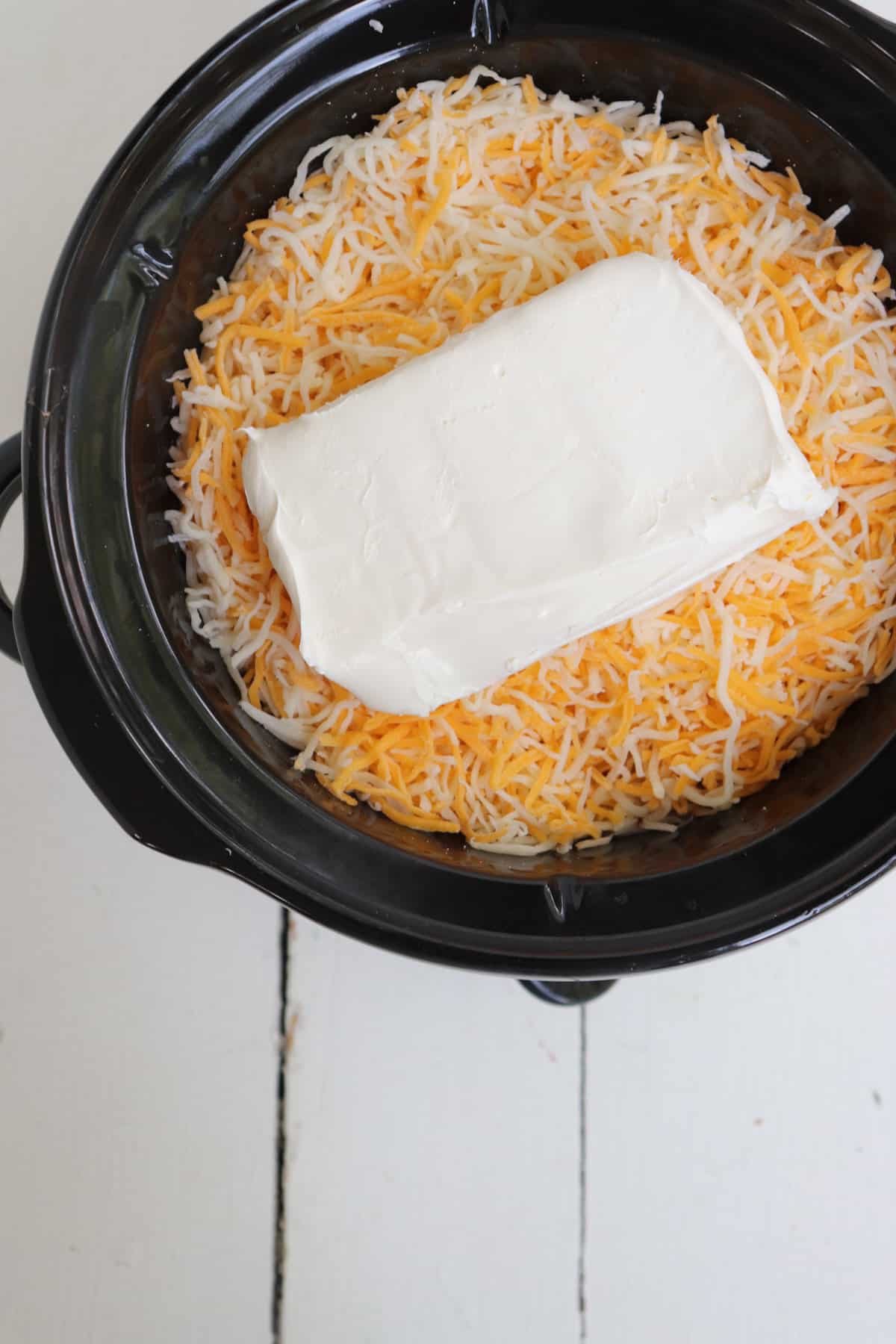 block of cream cheese on top of shredded cheese in crock pot.