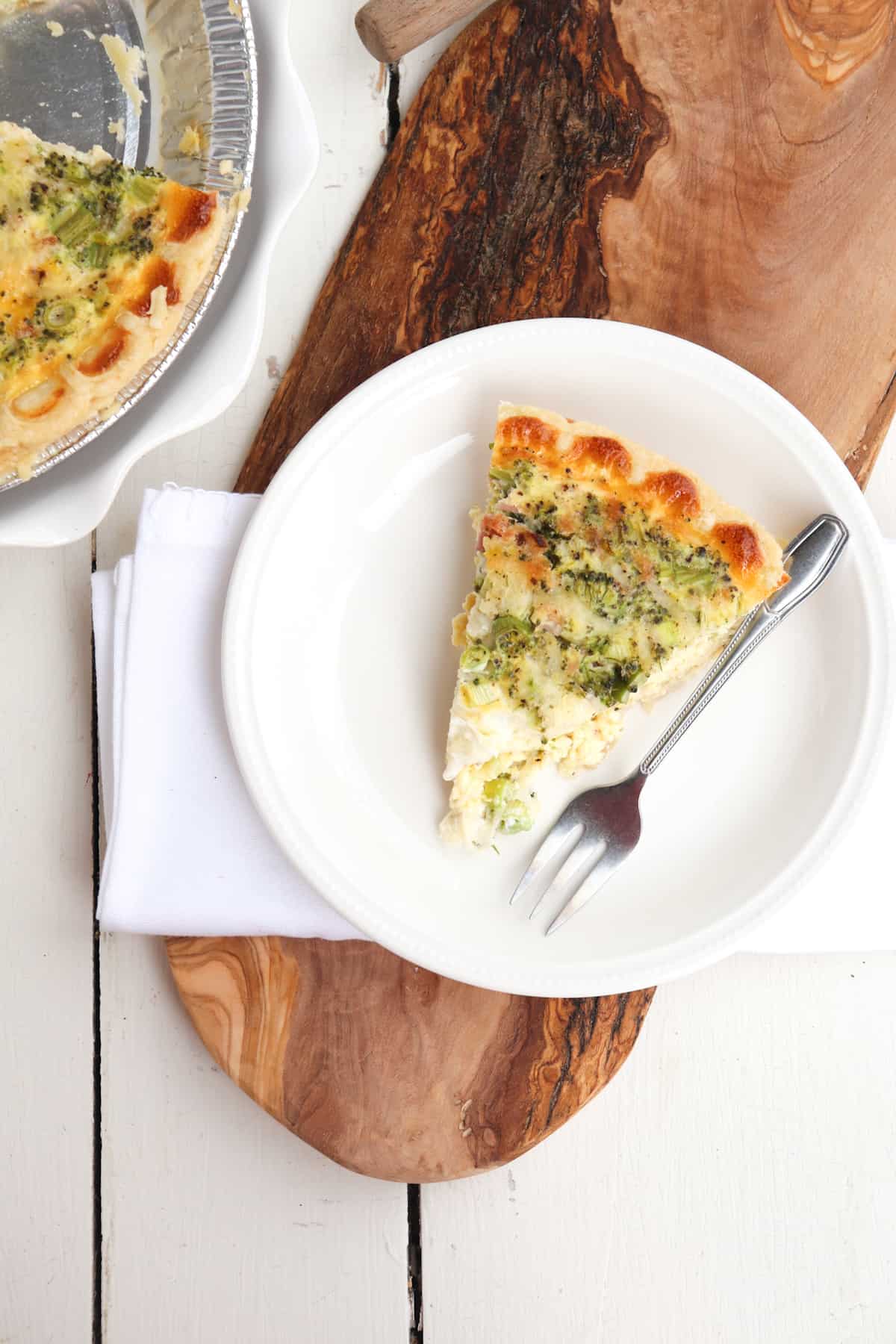 slice of ham and broccoli quiche on a white plate with whole quiche to the side.