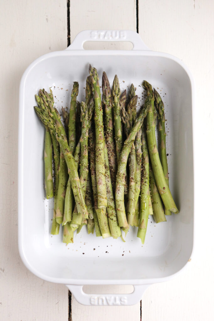 trimmed asparagus spears in white baking dish with pepper on top.