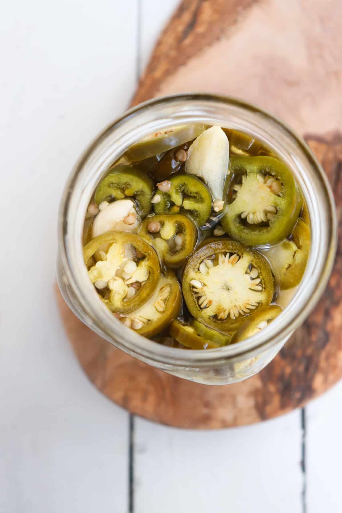 farther away overhead shot of finished fermented jalapenos in a glass jar on a wooden board.