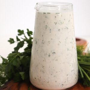 homemade ranch dressing in a glass container on a wooden board with herbs around it.