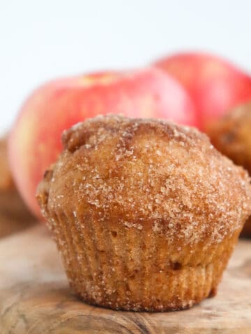 one apple cider muffin on a wooden cutting board with apples in the background.