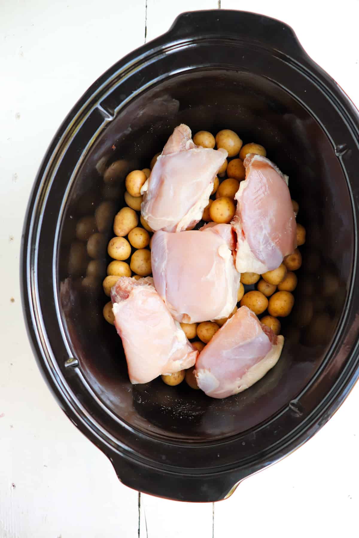 chicken thighs placed on layer of baby potatoes in a black slow cooker.