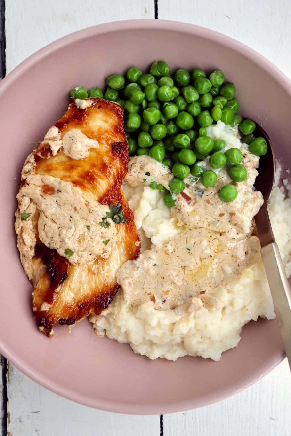browned chicken with boursin sauce, mashed potatoes, and peas in a pink bowl with a spoon.
