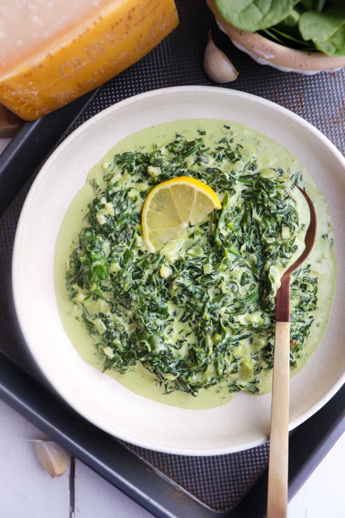 creamed spinach in a bowl topped with a lemon slice with a serving spoon and ingredients in the background.