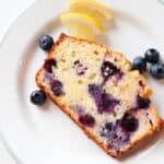 slice of lemon blueberry ricotta pound cake on a white plate with fruit scattered around.