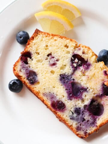 slice of lemon blueberry ricotta pound cake on a white plate with fruit scattered around.