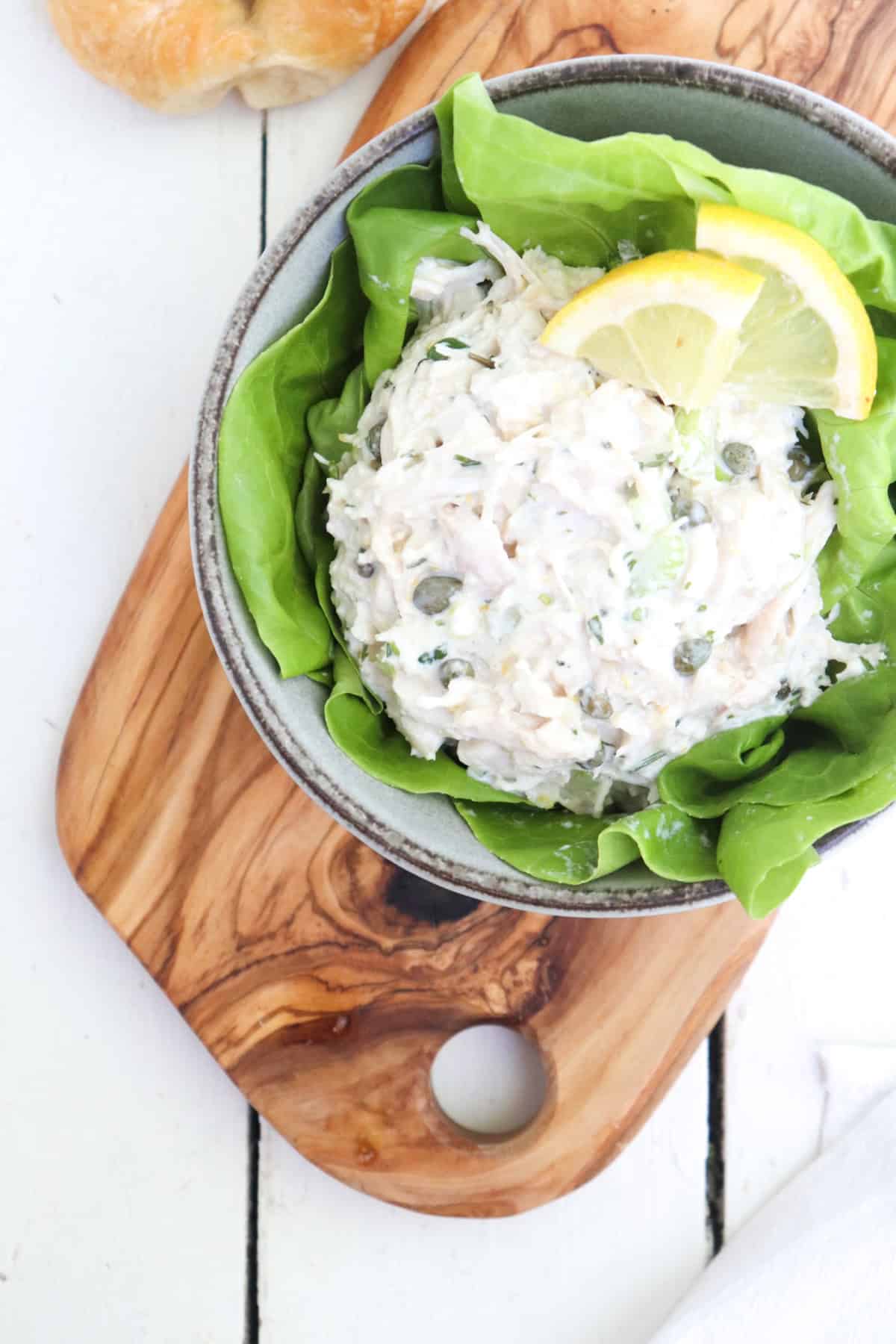 scoop of caper chicken salad in a bowl on a bed of lettuce with two small lemon wedges.