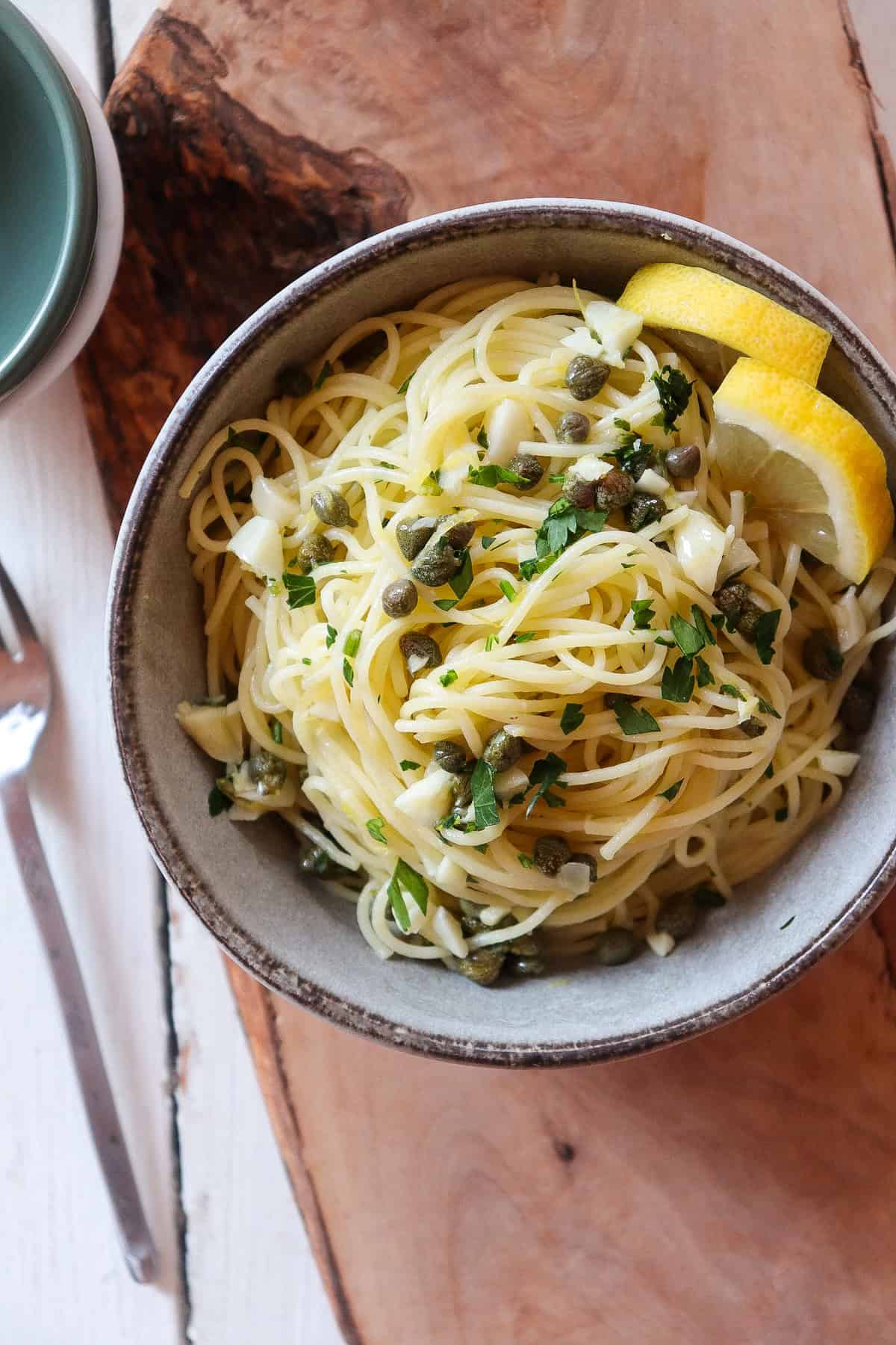 grey bowl of lemon caper pasta garnished with fresh lemons on a wooden board.