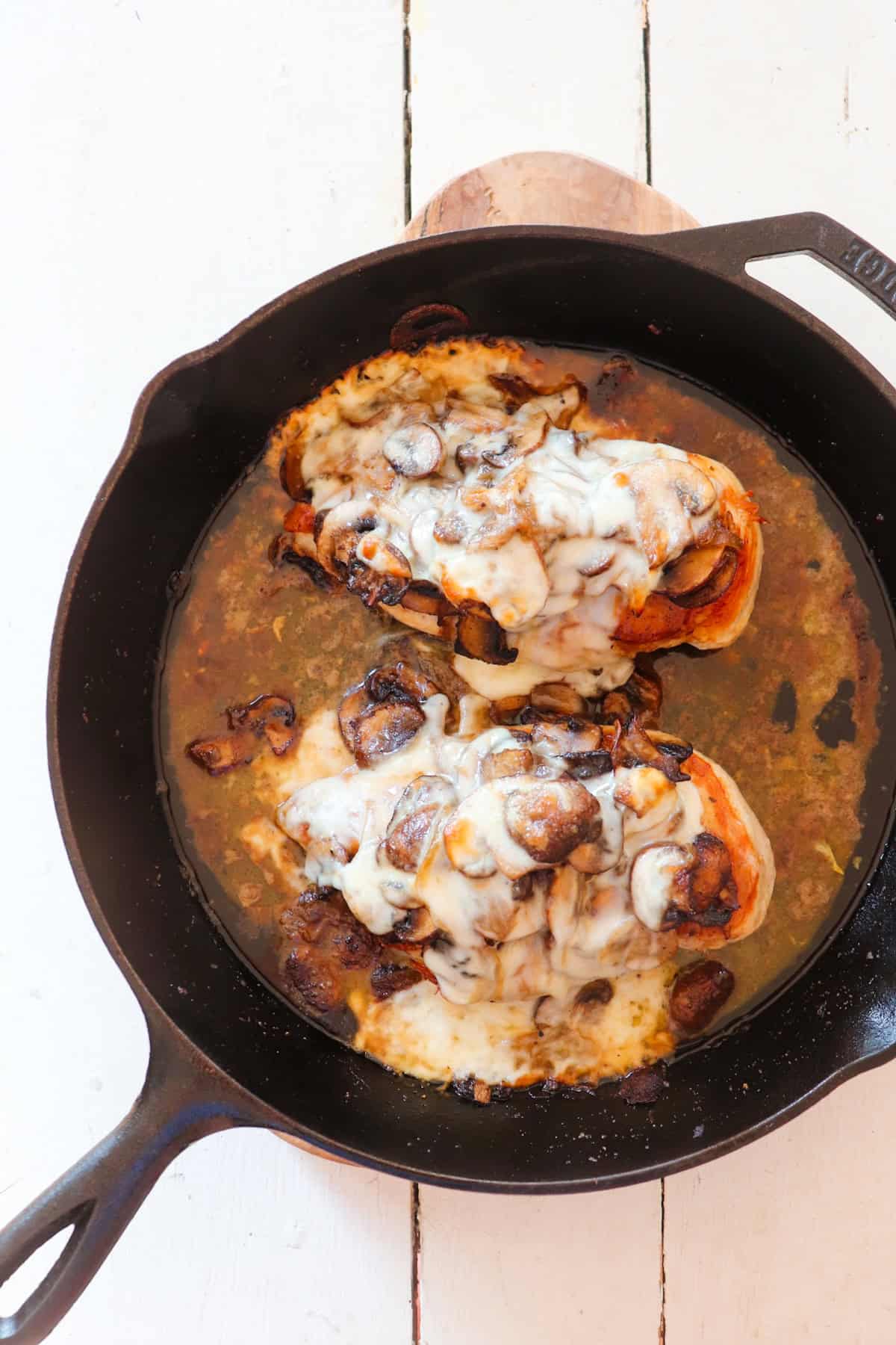 two chicken breasts smothered in mushrooms, onions, and melted cheese in a cast iron skillet.