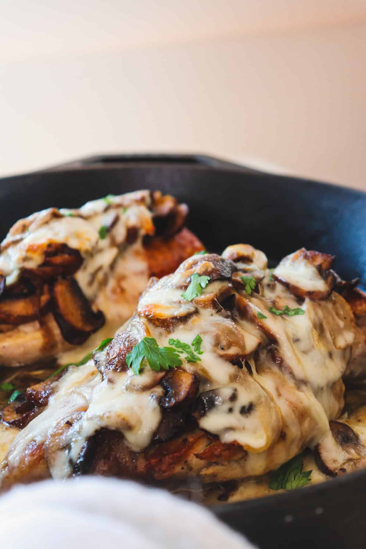 two chicken breasts smothered in mushrooms, onions, and melted cheese in a cast iron skillet with one shown closer.