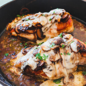 two chicken breasts smothered in mushrooms, onions, and melted cheese in a cast iron skillet.